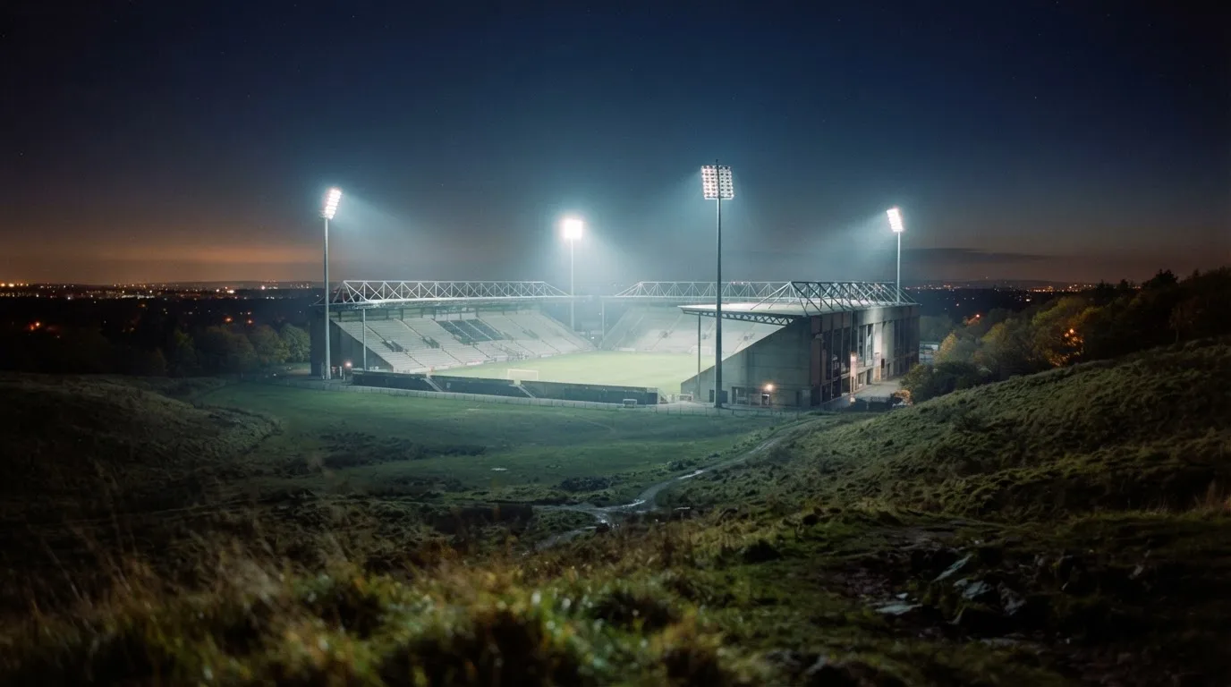 Ein Stadion unter Flutlicht bei Nacht, von einem Hügel aus der Ferne betrachtet, mit Dunst über dem Spielfeld.