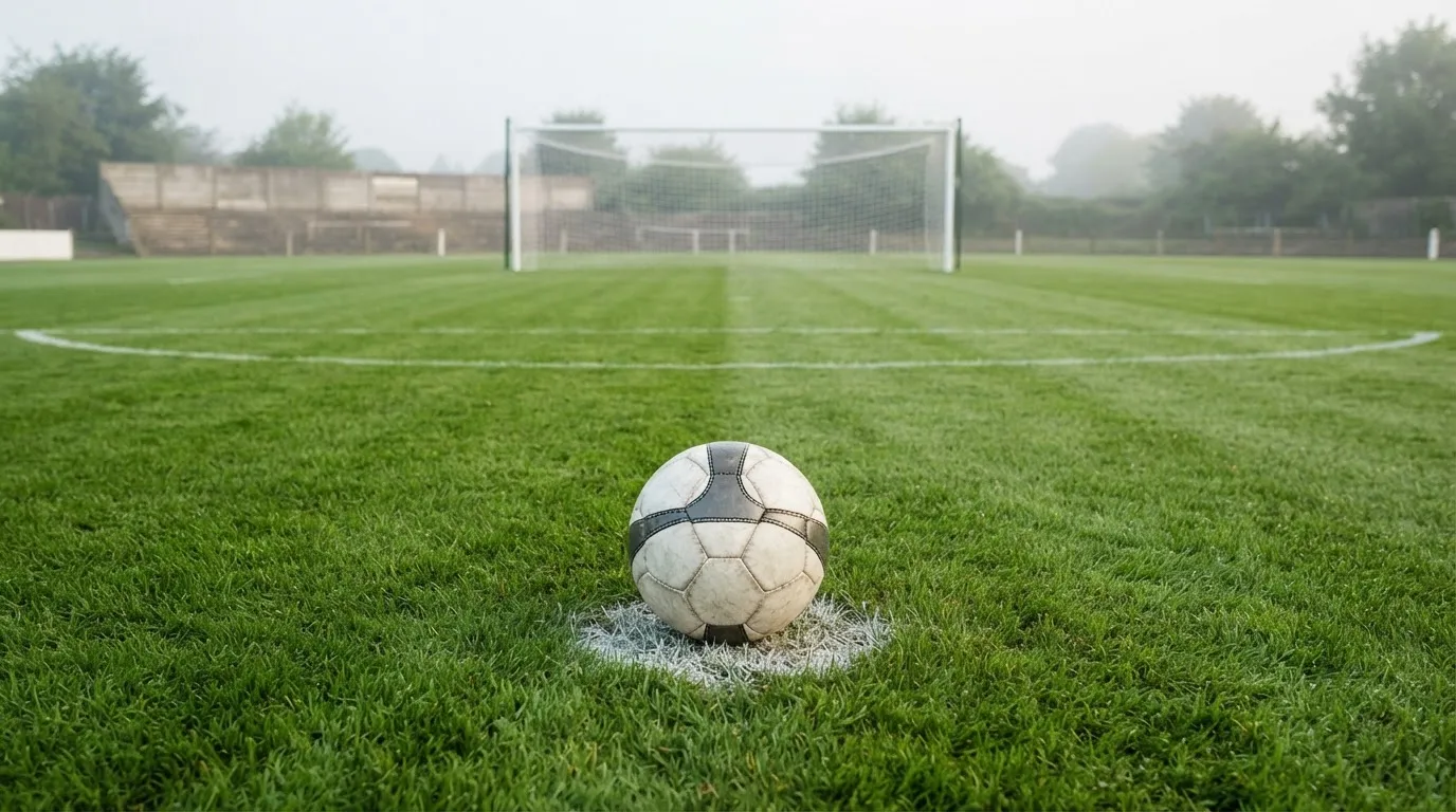 Ein Fußball liegt auf dem Elfmeterpunkt eines leeren Rasenplatzes, im Hintergrund eine Tribüne im Dunst.