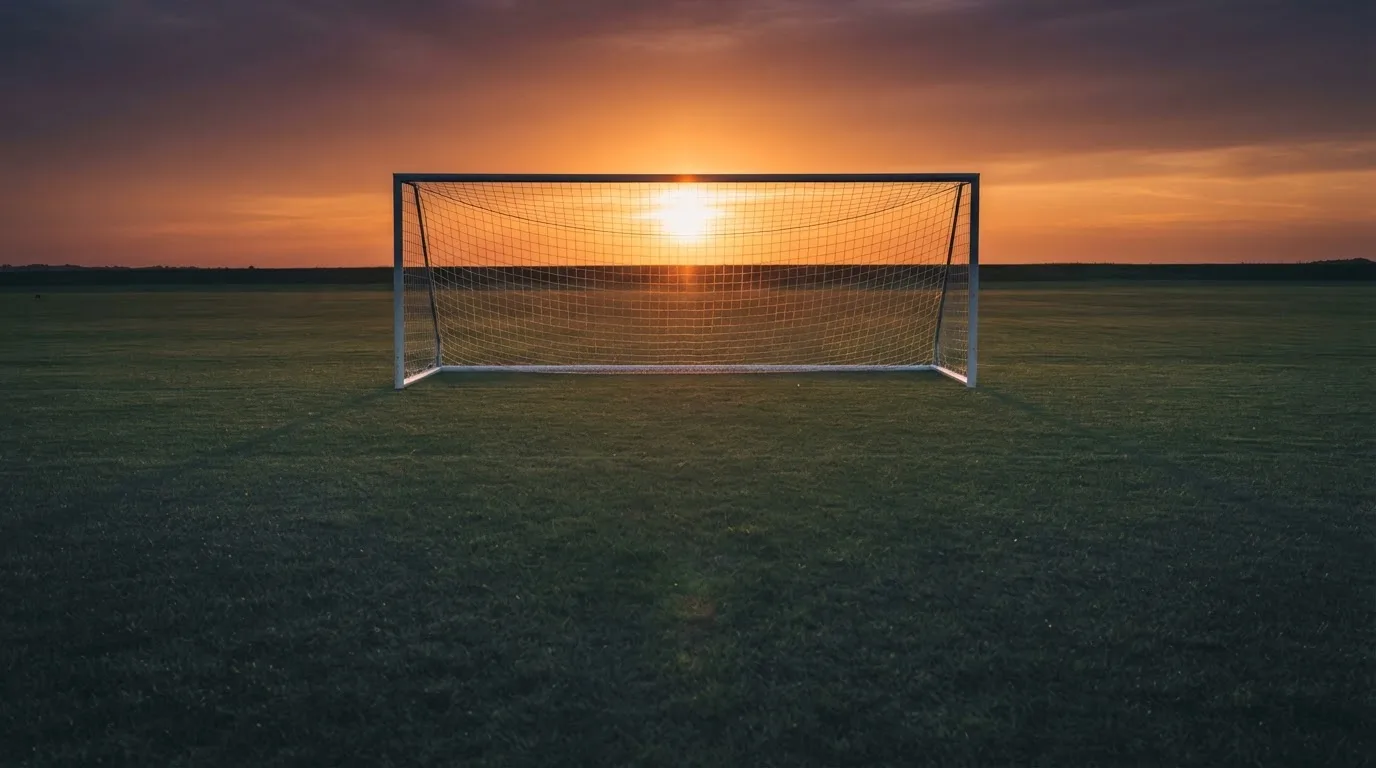 Ein Fußballtor mit Netz bei Sonnenuntergang auf einem leeren Platz, die Schatten der Torpfosten liegen lang auf dem Gras.