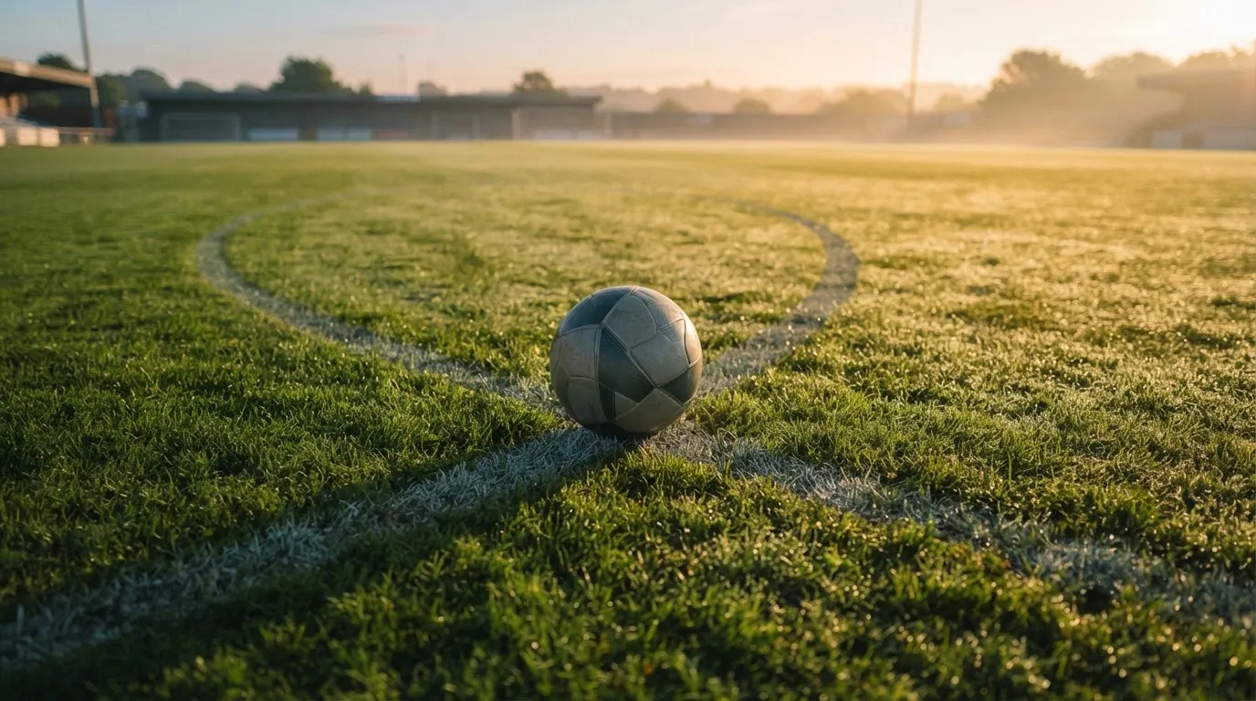 Ein Fußball liegt auf dem Anstoßpunkt eines Rasens bei Sonnenaufgang, Tau glitzert auf den Halmen.