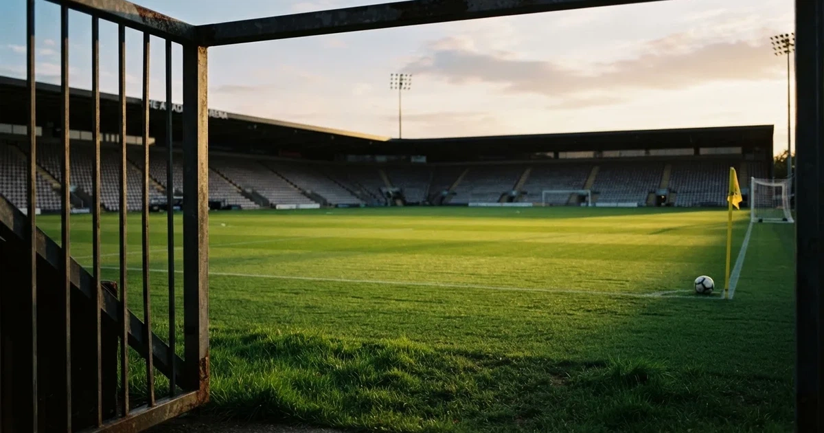 Blick durch ein Stadiontor auf einen grünen Rasen im Abendlicht, ein Ball ruht an der Eckfahne, die Tribünen sind leer.