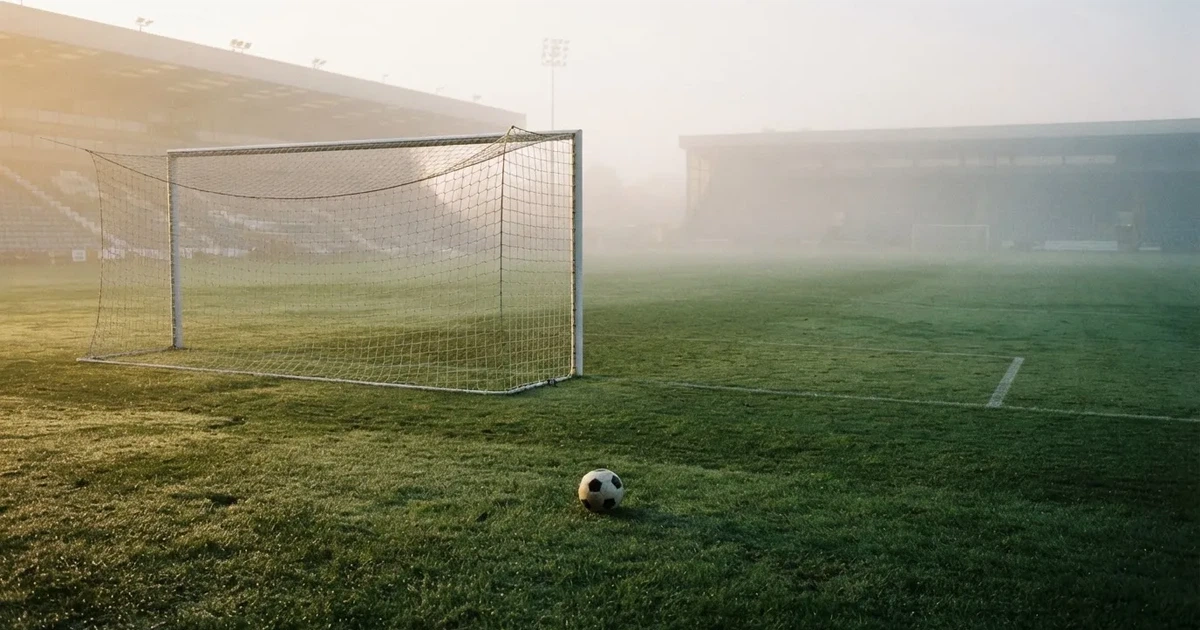 Ein Fußballtor mit gespanntem Netz steht auf einem leeren Rasenplatz, davor ein einzelner Ball, Morgendunst liegt über dem Feld.