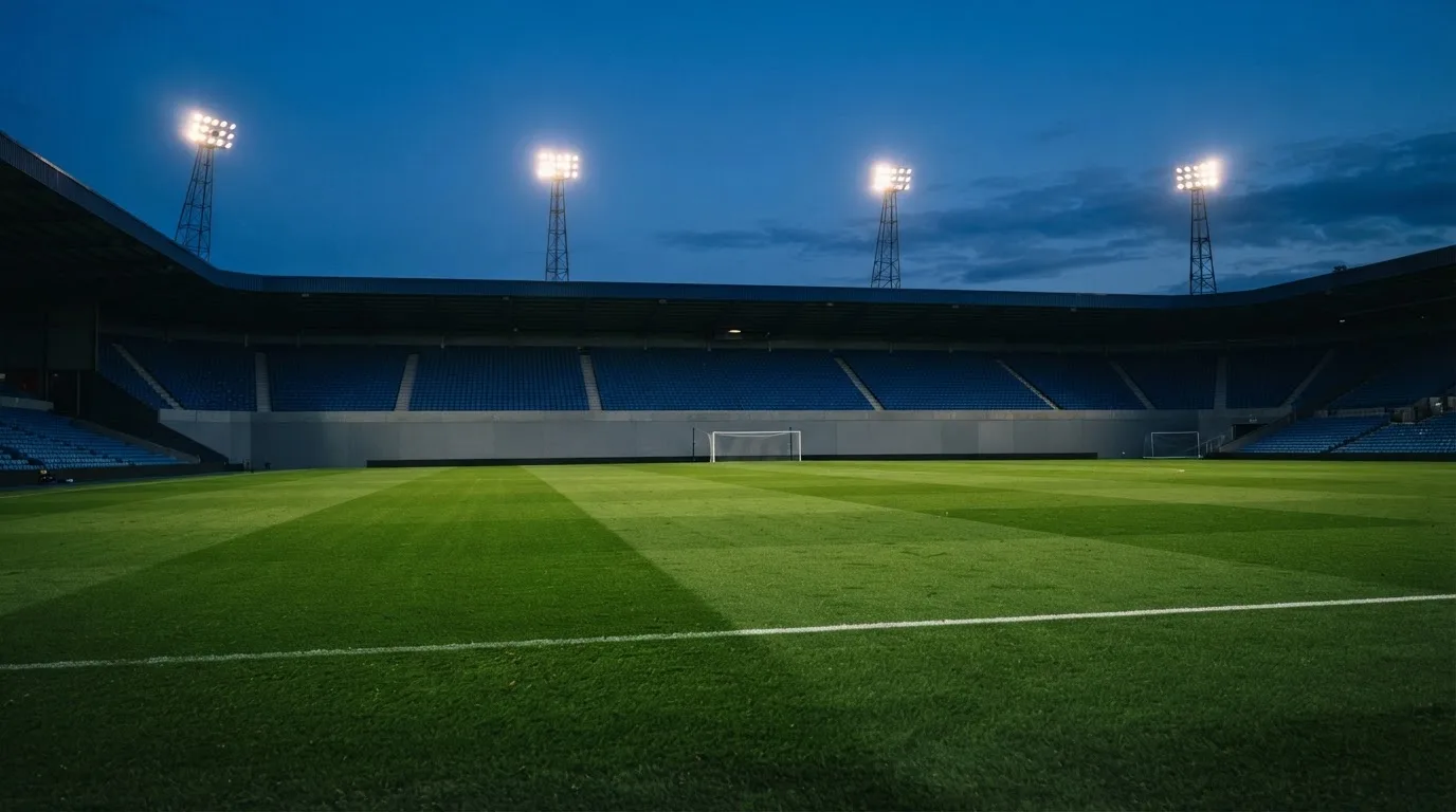 Eine leere Stadiontribüne unter Flutlicht aus der Ferne betrachtet, die Rasenfläche leuchtet grün in der Dämmerung.
