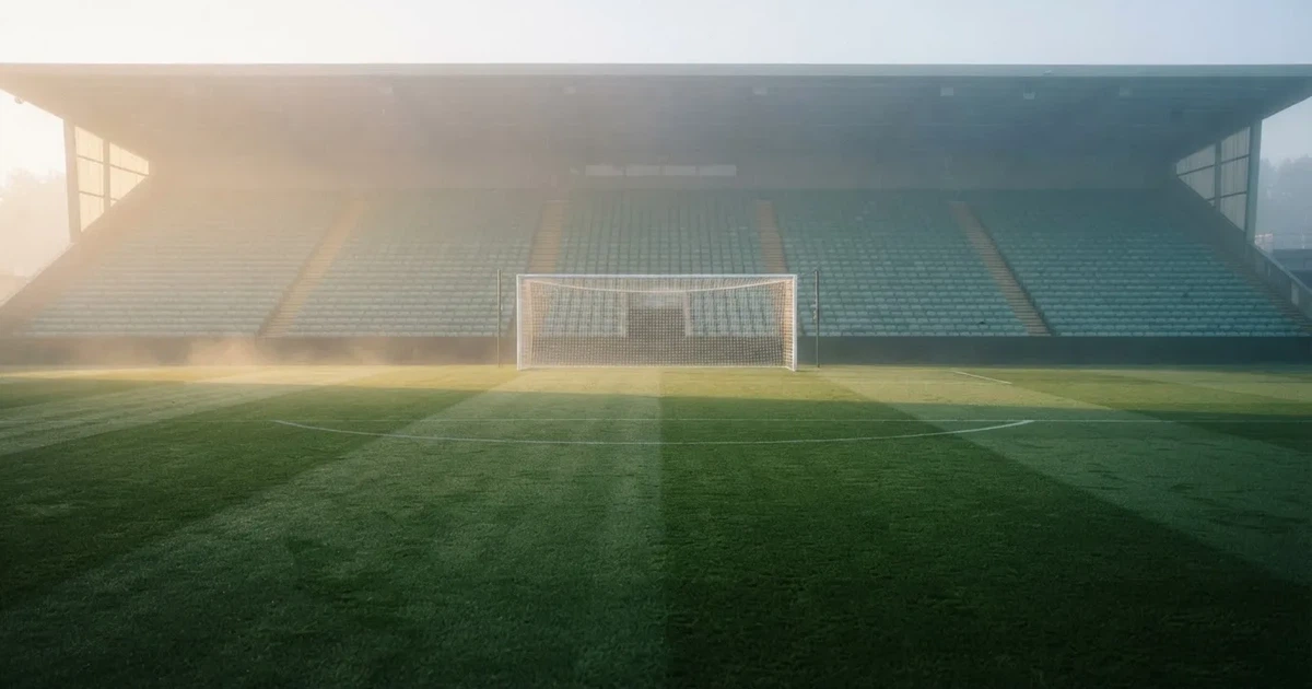 Ein Fußballplatz mit gepflegtem Rasen und einem Tor, über dem Spielfeld liegt Morgennebel, im Hintergrund eine leere Tribüne.
