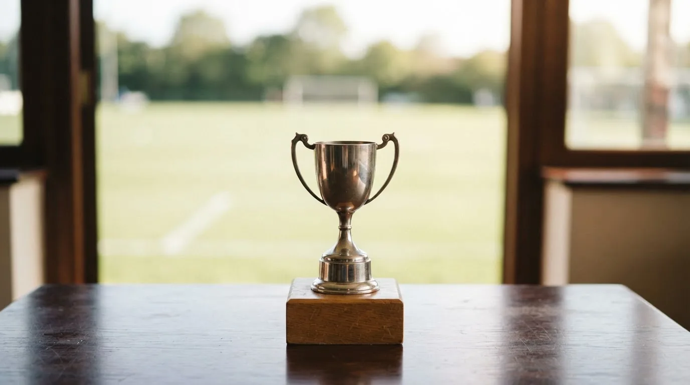 Ein kleiner Pokal aus Metall ohne Gravur steht auf einem Podest aus Holz, dahinter unscharf ein Fußballfeld.