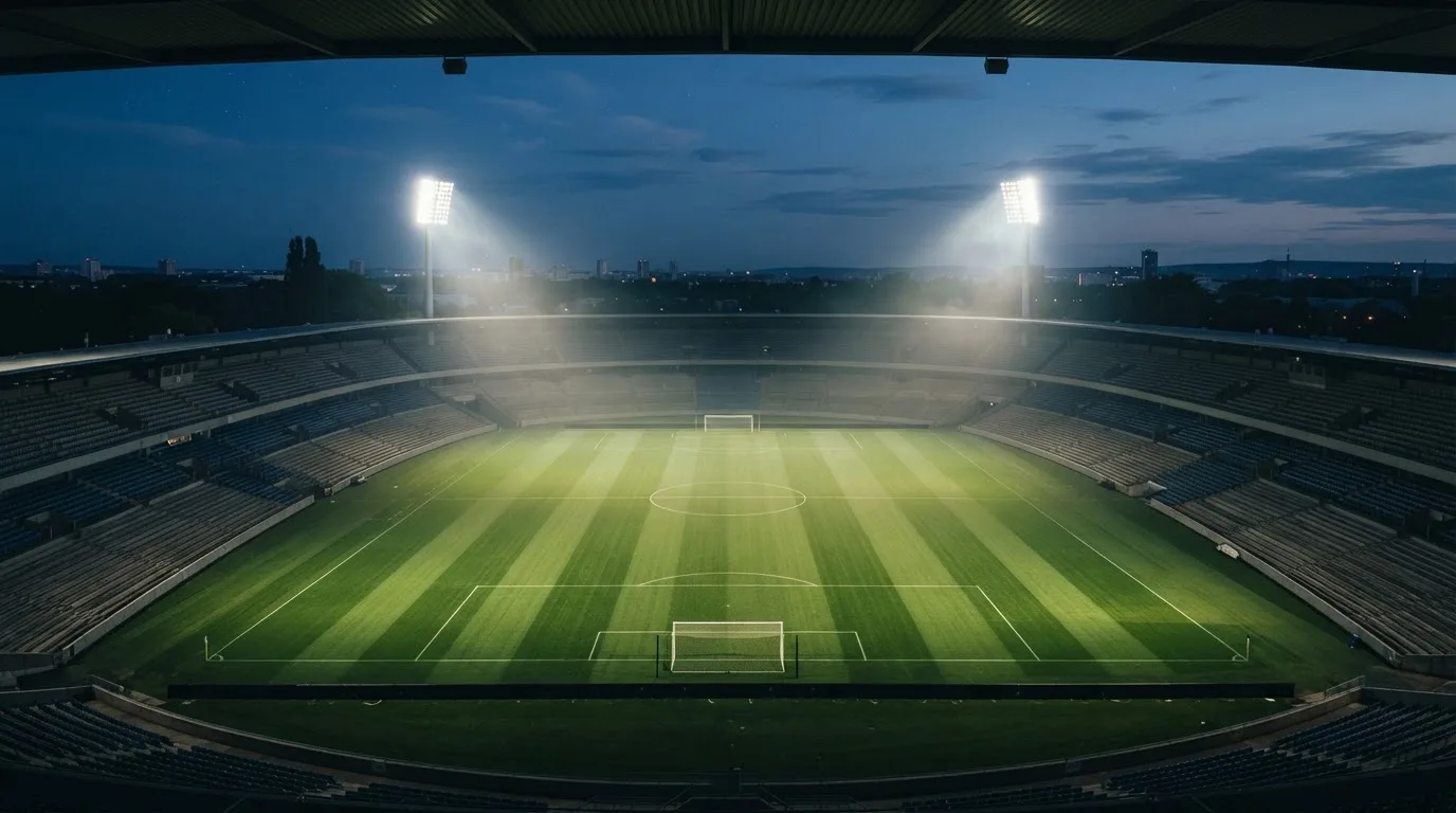 Eine leere Stadiontribüne unter Flutlicht aus der Ferne betrachtet, die Rasenfläche leuchtet grün in der Dämmerung.